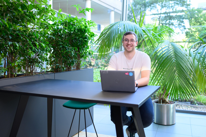 Ein junger lächelnder Mann sitzt an einem Tisch, auf dem sein Laptop steht. Im Hintergrund stehen große grüne Pflanzen.